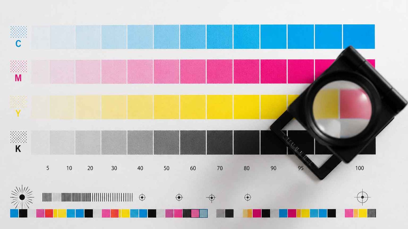 A printed CMYK control strip showing graduated cyan, magenta, yellow and black patches, with a printer&rsquo;s loupe resting on the sheet